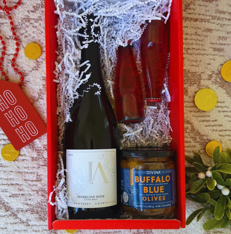 A red holiday box holding AJA sparkling wine, bitter soda, and a jar of blue cheese olives